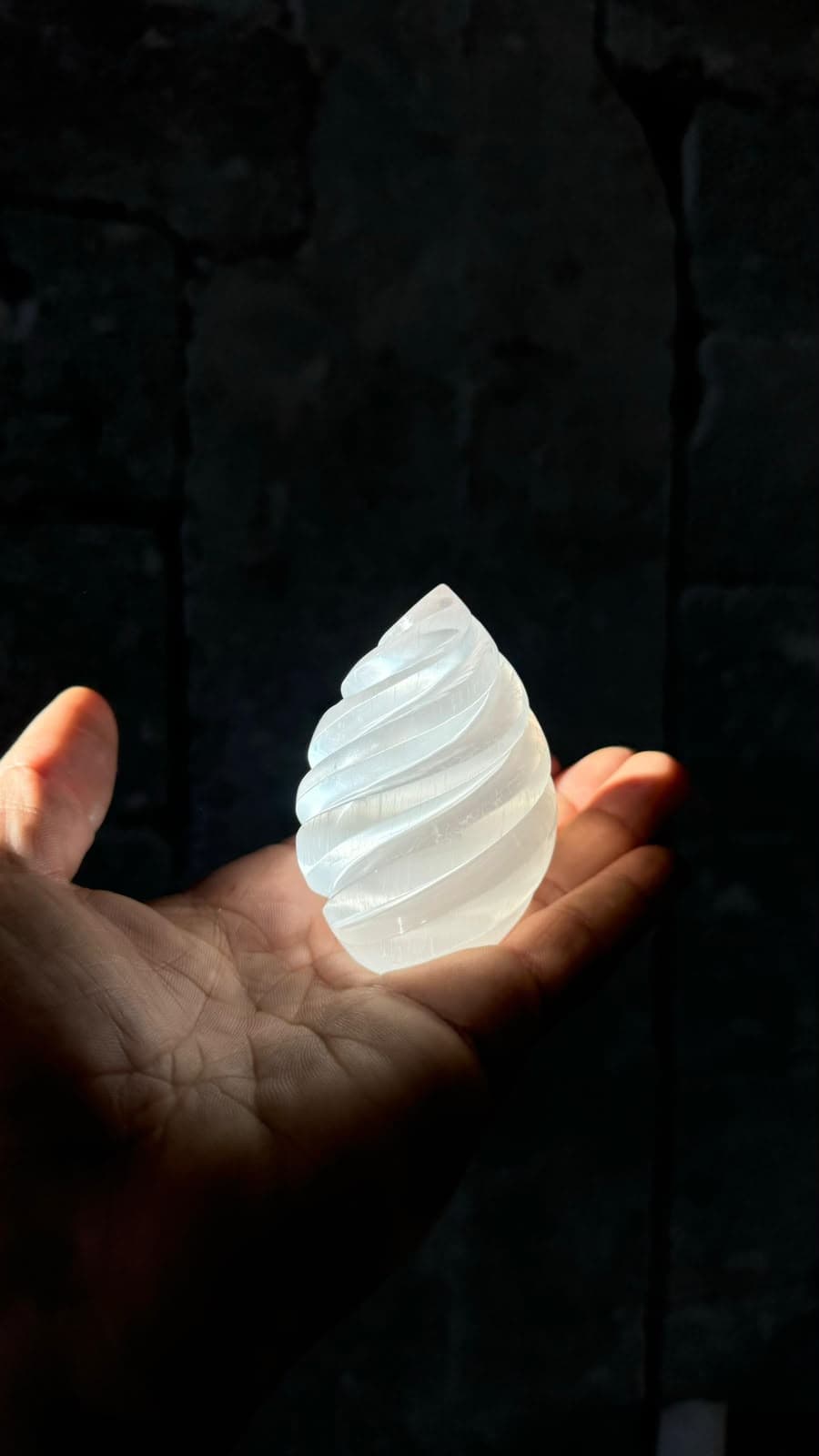 Hand holding a glowing white spiral selenite crystal against a dark, shadowy stone background.